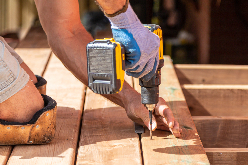 A close up of an electric drill being used to screw in a plank of wood. You can see several wood planks to the left of the drill and a set of arms operating it. This man is installing a deck.