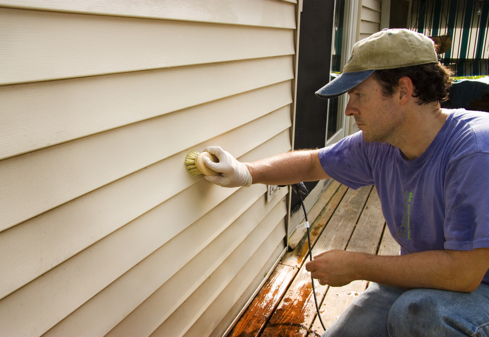 A worker in a blue uniform and baseball cap uses his arm to press a yellow sponge against a house's white siding. This is one of the recommended ways to clean your fiber cement siding.