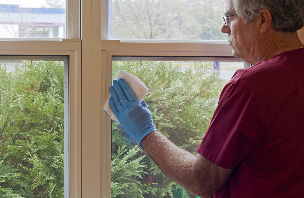 A man in a red shirt and blue gloves uses a white cloth to clean a window. You can see shrubs on the other side of the glass.